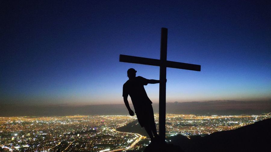 Man holding cross on cliff against illuminated city at night