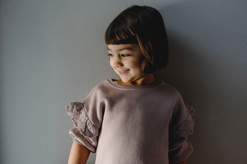 Cute little girl in casual outfit leaning on gray wall at home while smiling sincerely and looking away