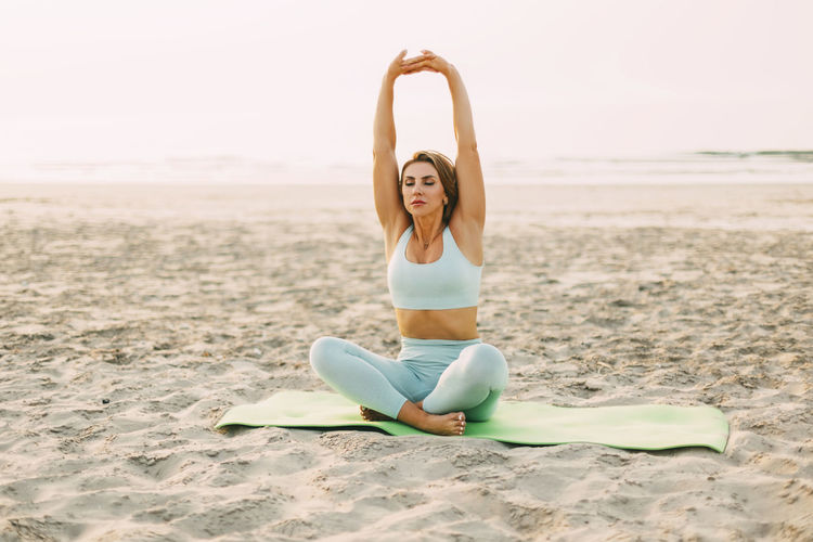An attractive girl is sitting in a lotus position and doing exercises on the beach at sunset. 