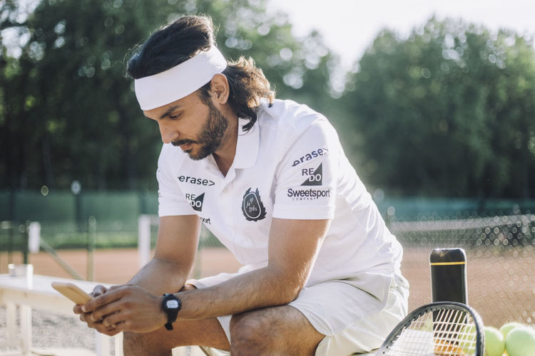 Man wearing headband using smart phone sitting on bench at tennis court