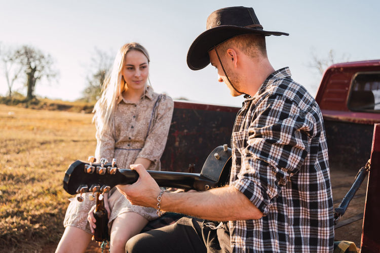 Boyfriend in cowboy hat playing acoustic guitar while sitting with girlfriend in trunk of red retro pickup car parked on sandy road in countryside