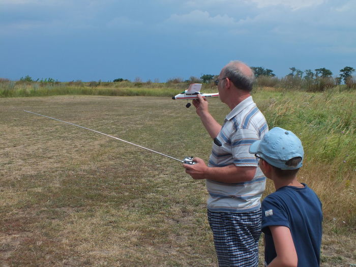 Grandfather holding remote controlled airplane with grandson on grassy field against cloudy sky