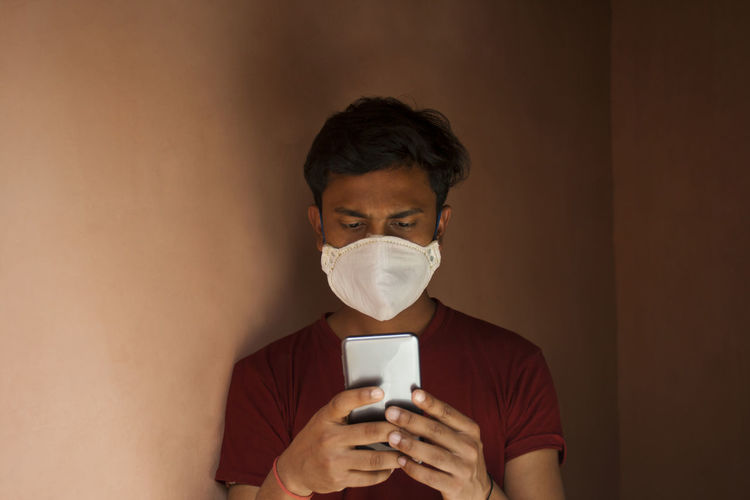 Close-up of young man wearing flu mask using smart phone against wall at home